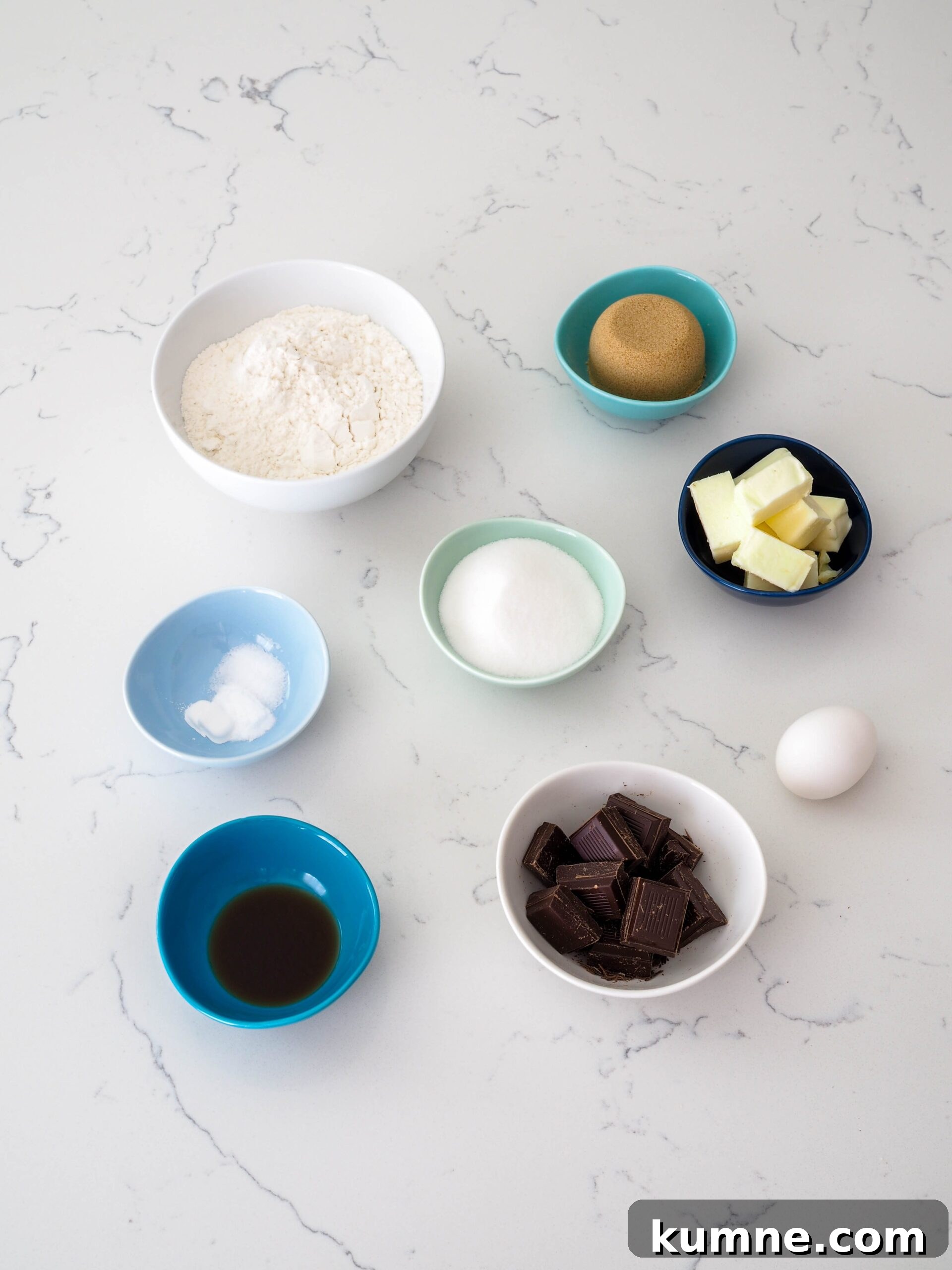 Decadent Dark Chocolate Chunk Cookie Recipe 3 Flour, sugar, brown sugar, butter, an egg, chocolate chunks, vanilla extract, baking powder and soda, and salt on a counter.