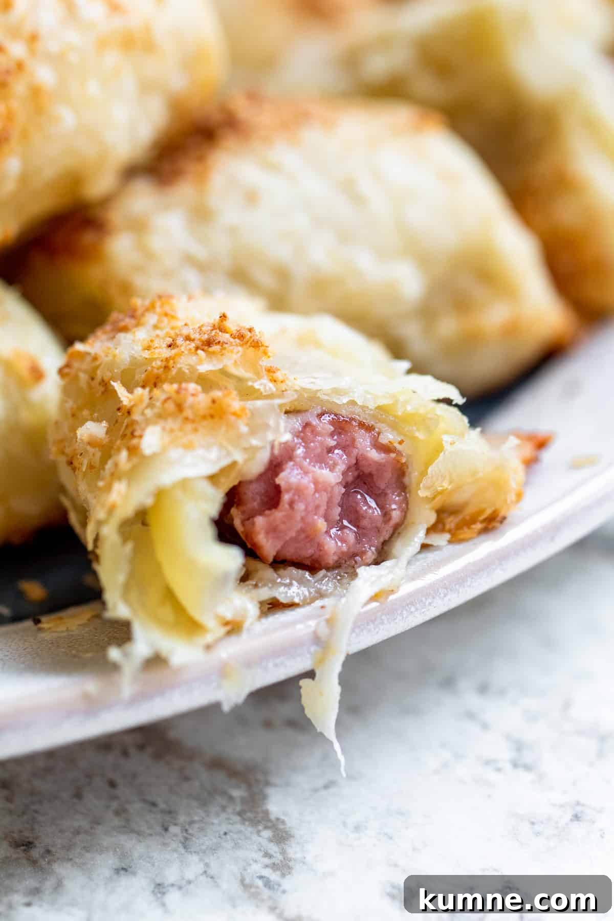 Cheesy Puff Pastry Pigs in a Blanket Recipe 6 Strait on shot of cheesy pigs in a blanket with bit out of it on plate.