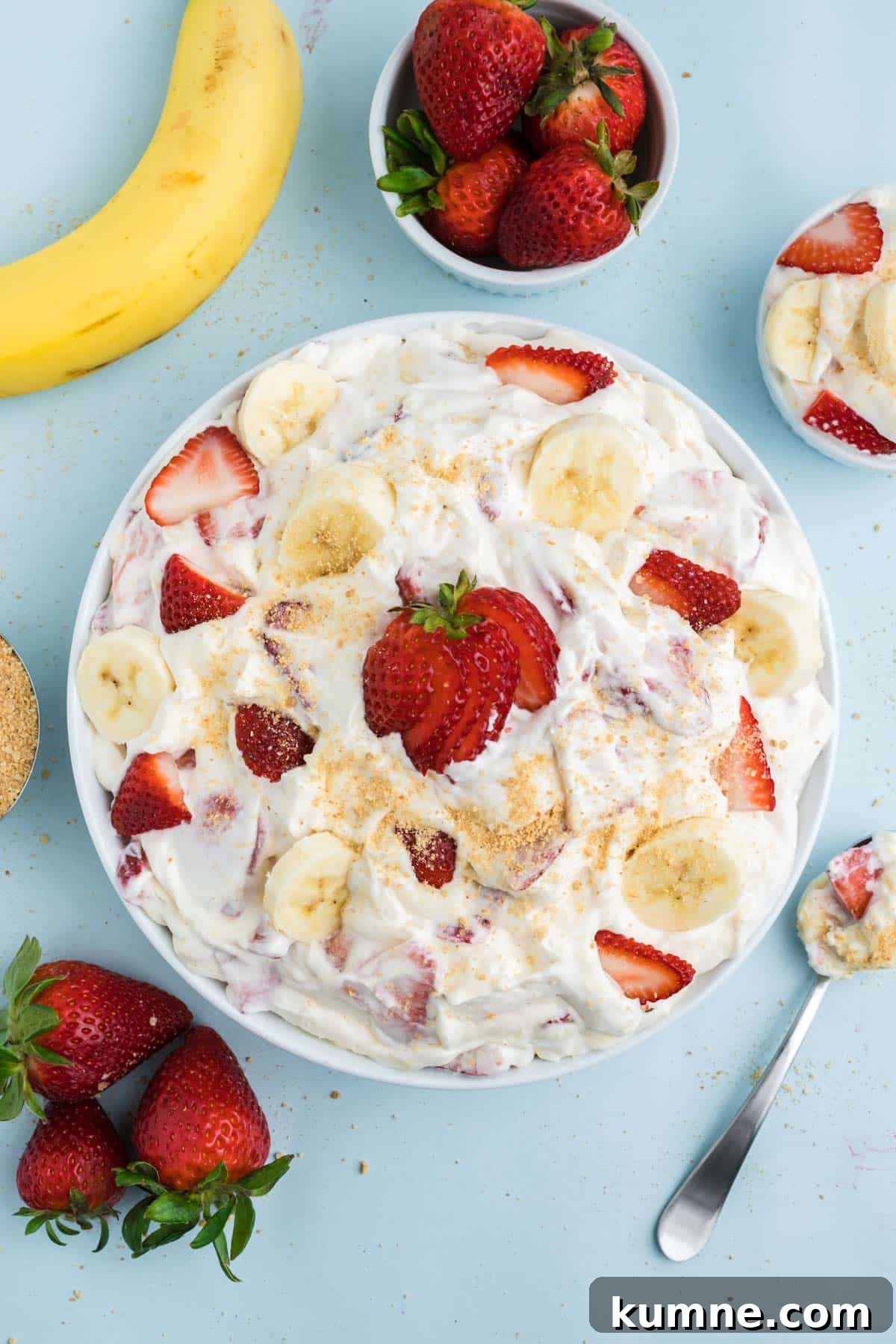 A bowl of strawberry banana cheesecake salad on the table with fresh fruit in bowls around it and a spoon to the side.