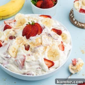 Strawberry banana cheesecake salad in a bowl on the table with a spoonful next to the bowl and fresh fruit around it.