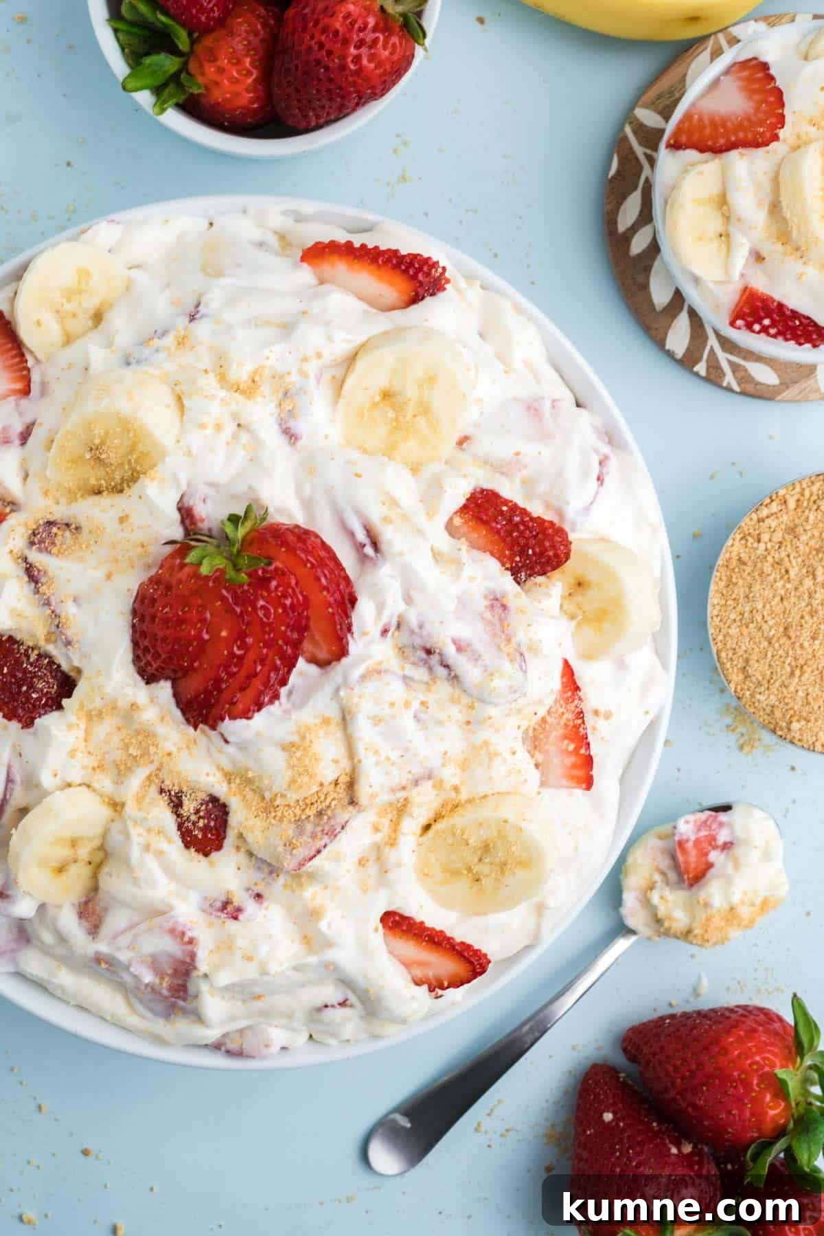 A bowl of cheesecake fruit salad on the table with a fresh fruit and a spoonful next to the bowl.