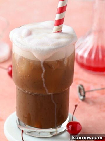 A cherry americola drink in a clear glass with a straw on a pink surface.
