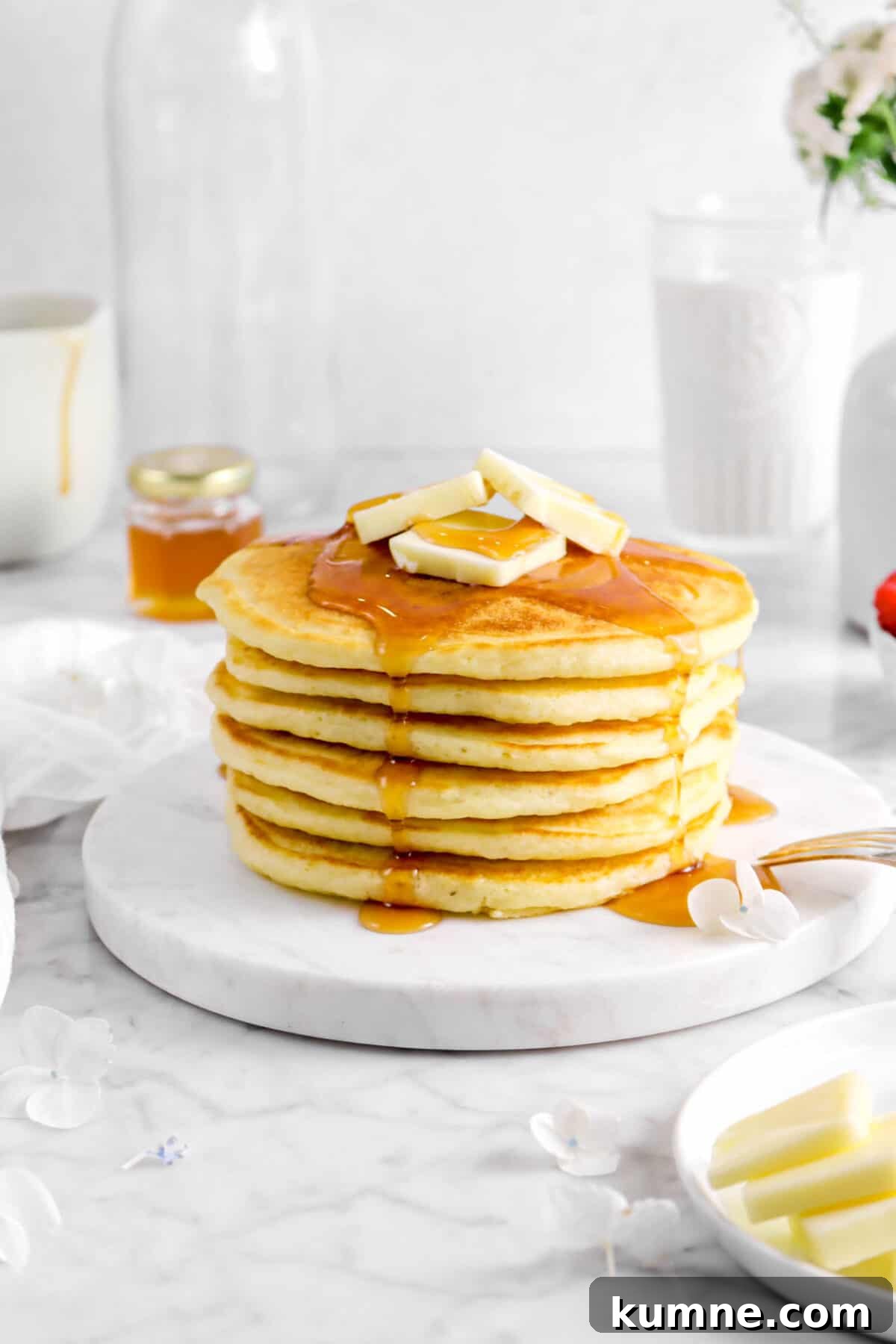 Fluffy Homemade Buttermilk Pancakes Recipe for Perfect Mornings 10 pancakes on marble plate with syrup and butter, flowers around, and glass of milk