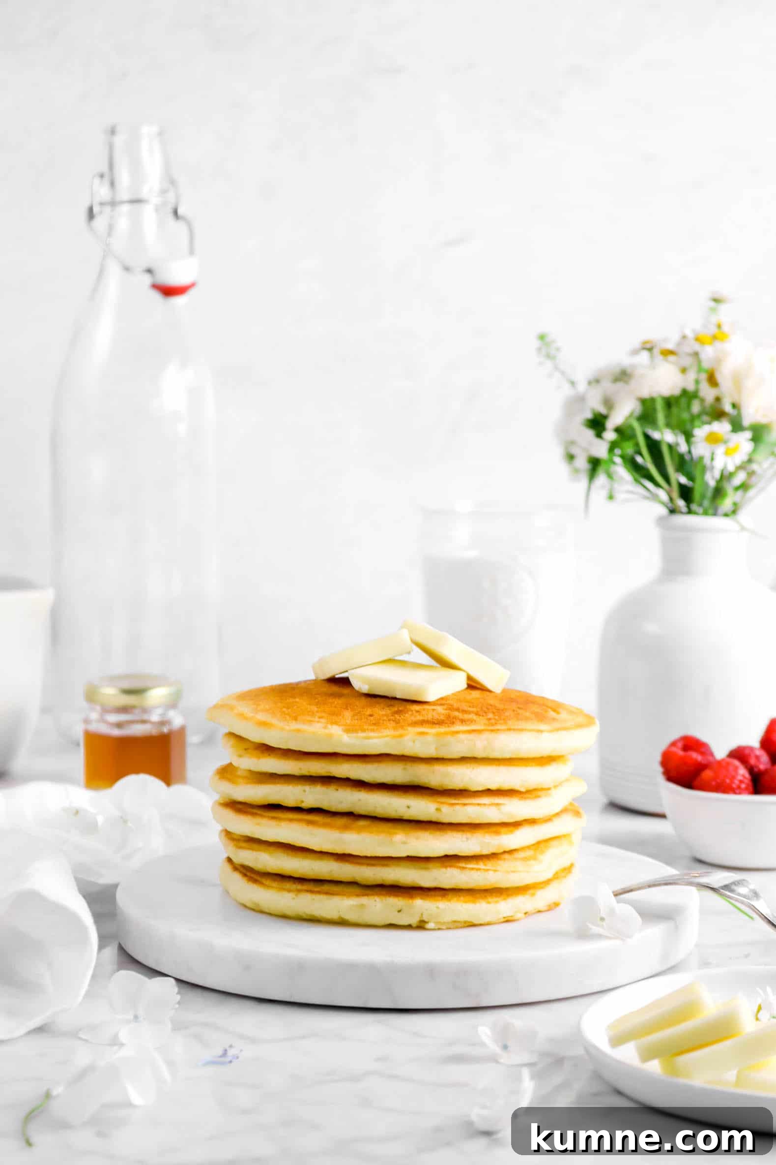 Fluffy Homemade Buttermilk Pancakes Recipe for Perfect Mornings 4 stack of pancakes on marble plate with overturned fork, sliced butter on white plate, raspberries in white bowl, flowers in vase, jar of honey, glass of milk, empty bottle, white napkin