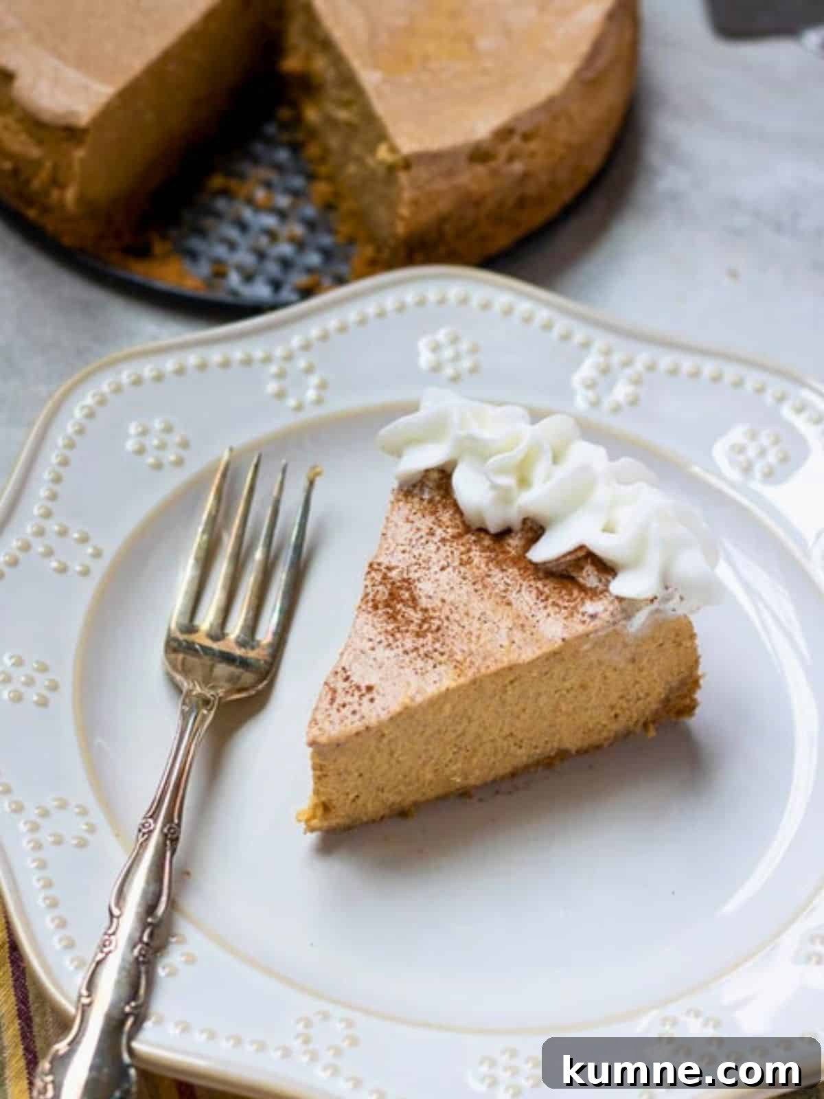 Instant Pot Pumpkin Cheesecake Slice of Instant Pot Pumpkin Cheesecake on plate with homemade whipped cream and fork with remaining pumpkin cheesecake in background.