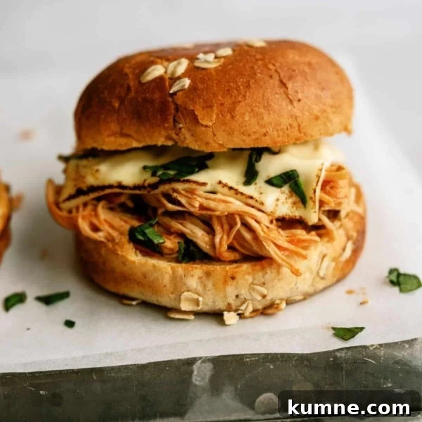 10 Make-Ahead Slow Cooker Freezer Meals You Can Prep in 90 Minutes 9 A sandwich with shredded chicken, melted cheese, and greens in a sesame seed bun, placed on white parchment paper.
