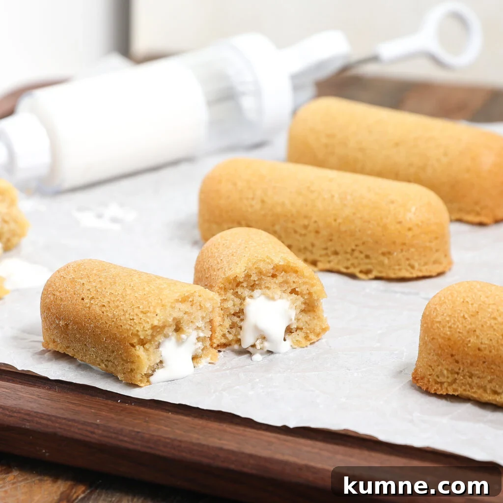 Golden sponge cakes filled with white cream are arranged on parchment paper, with one homemade Twinkie split open to show the creamy filling. A cream-filled syringe is in the background.