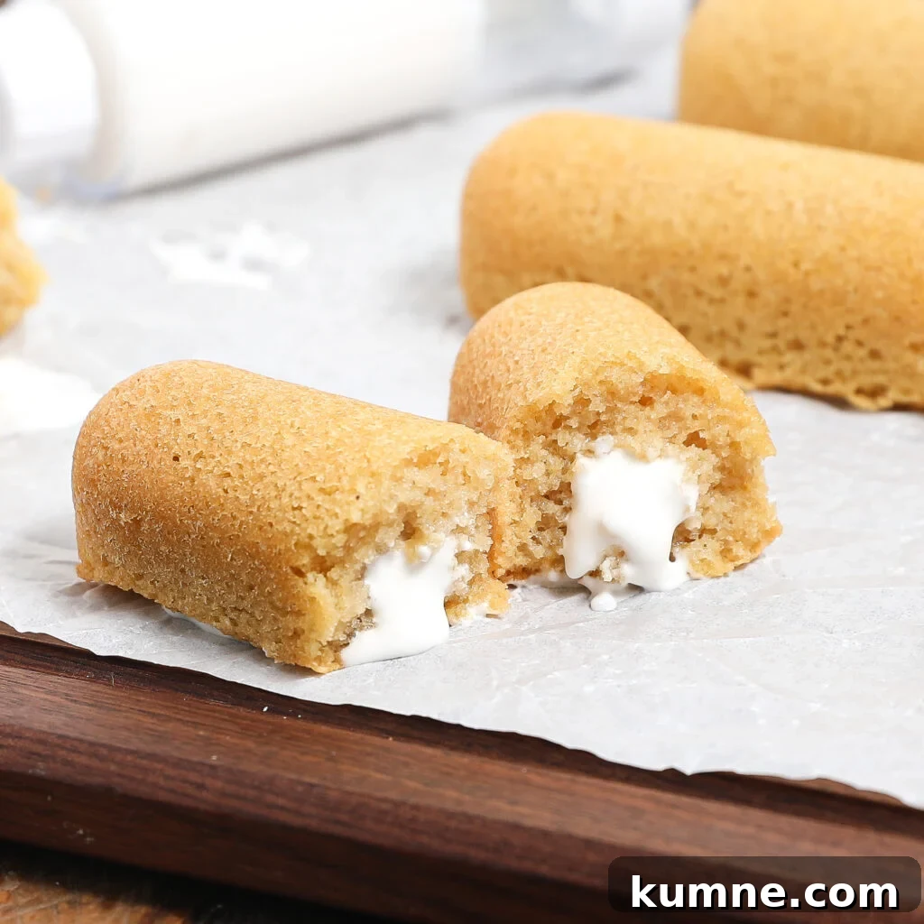 Two golden homemade Twinkies rest on parchment paper, one split open to reveal creamy white filling inside.
