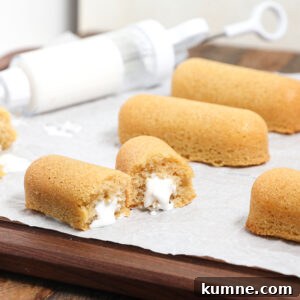 Golden sponge cakes filled with white cream are arranged on parchment paper, with one homemade Twinkie split open to show the creamy filling. A cream-filled syringe is in the background.
