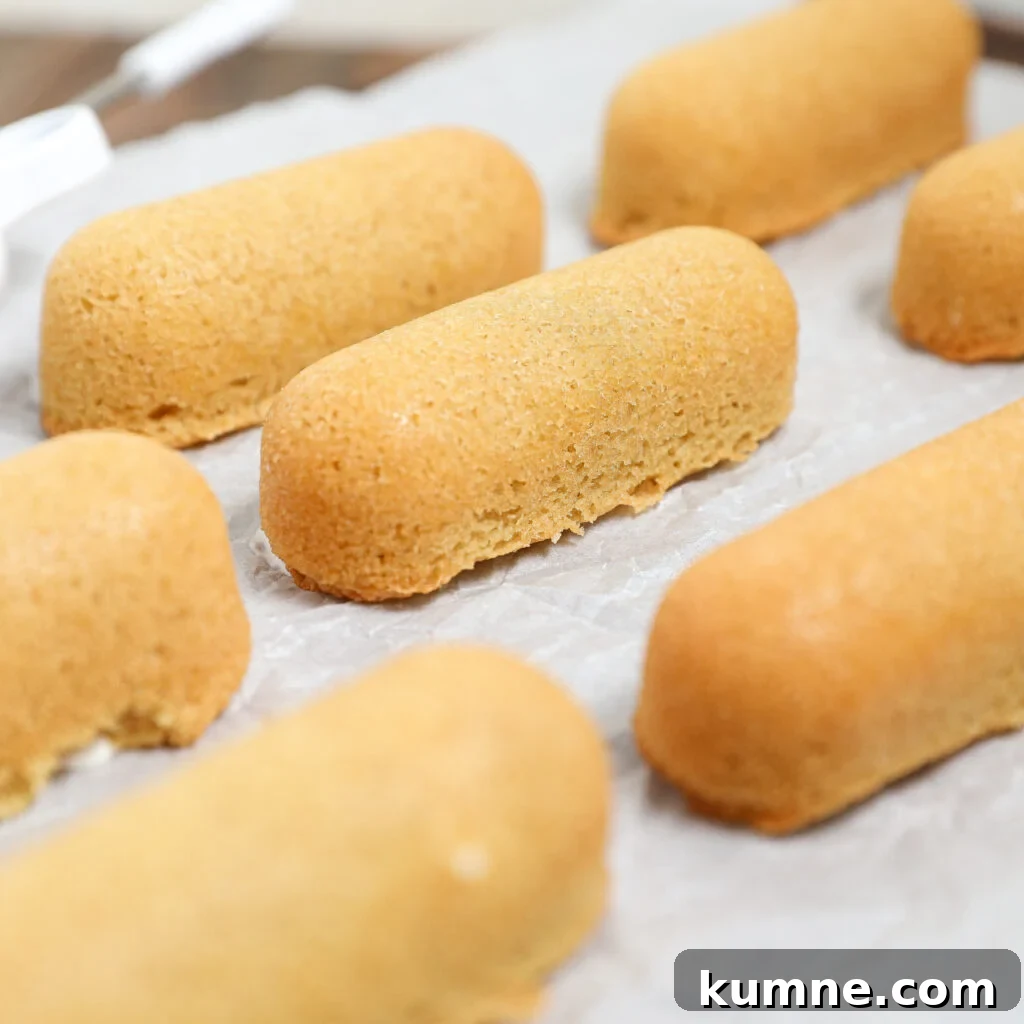 Golden brown sponge cakes, resembling homemade Twinkies, are arranged in rows on a sheet of parchment paper. The cakes have a smooth, slightly domed surface and appear soft and freshly baked.