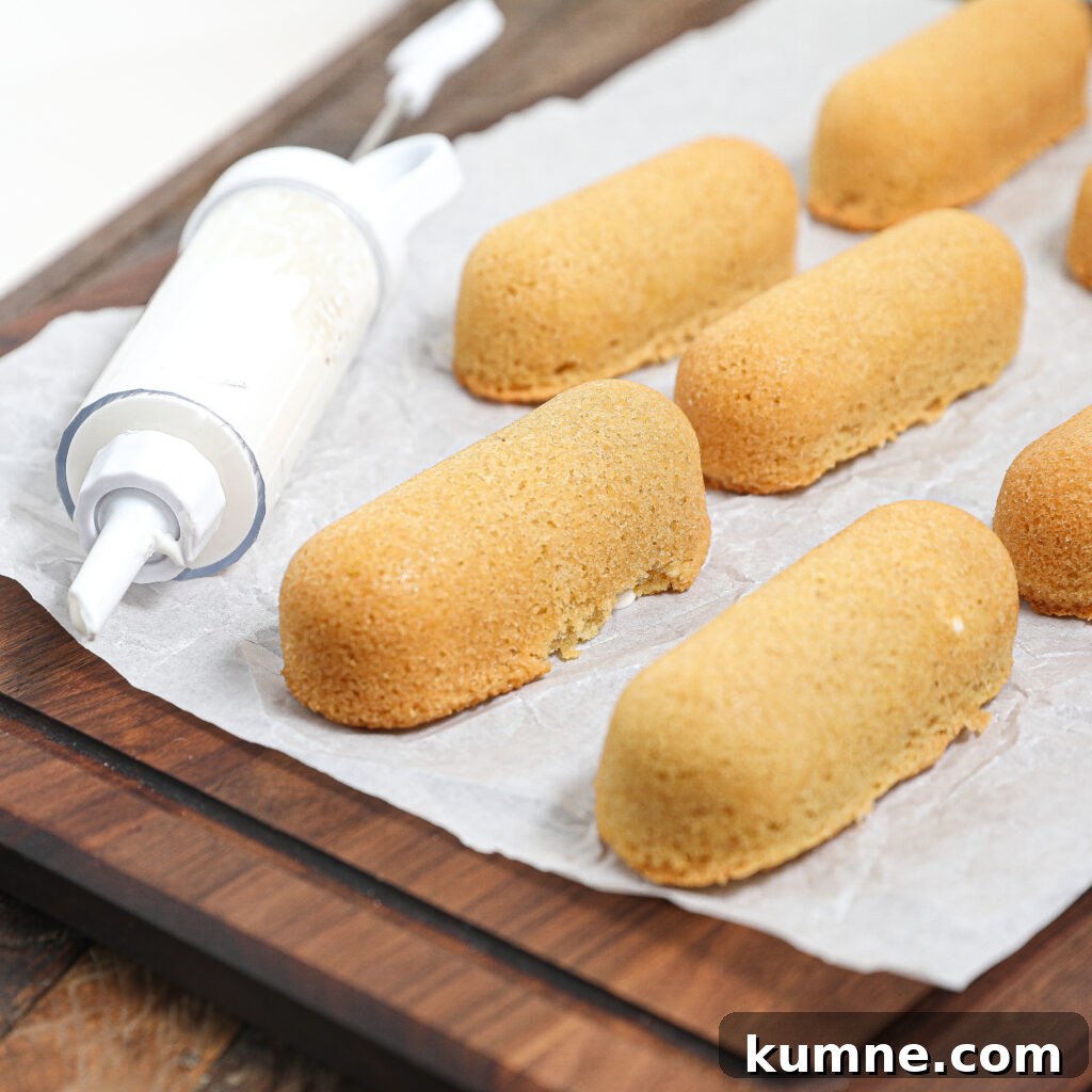 A piping tool filled with cream sits next to several golden homemade Twinkies on parchment paper atop a wooden board. One cake shows a small bite taken out of it.