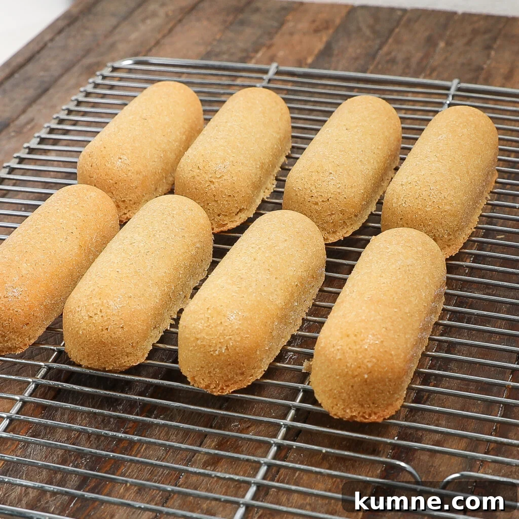 Eight golden-brown, oval homemade Twinkies cool on a metal wire rack placed on a wooden surface.