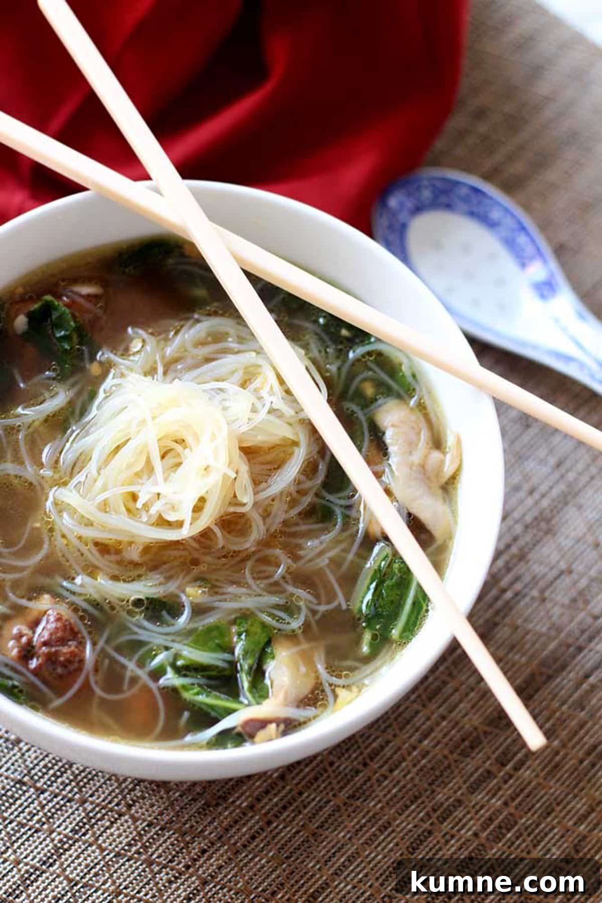 Authentic Asian Chicken Noodle Soup Recipe for Comforting Flavor 2 White bowl containing Asian Chicken Noodle Soup with crossed chopsticks sitting on a brown table mat, red napkin on table.