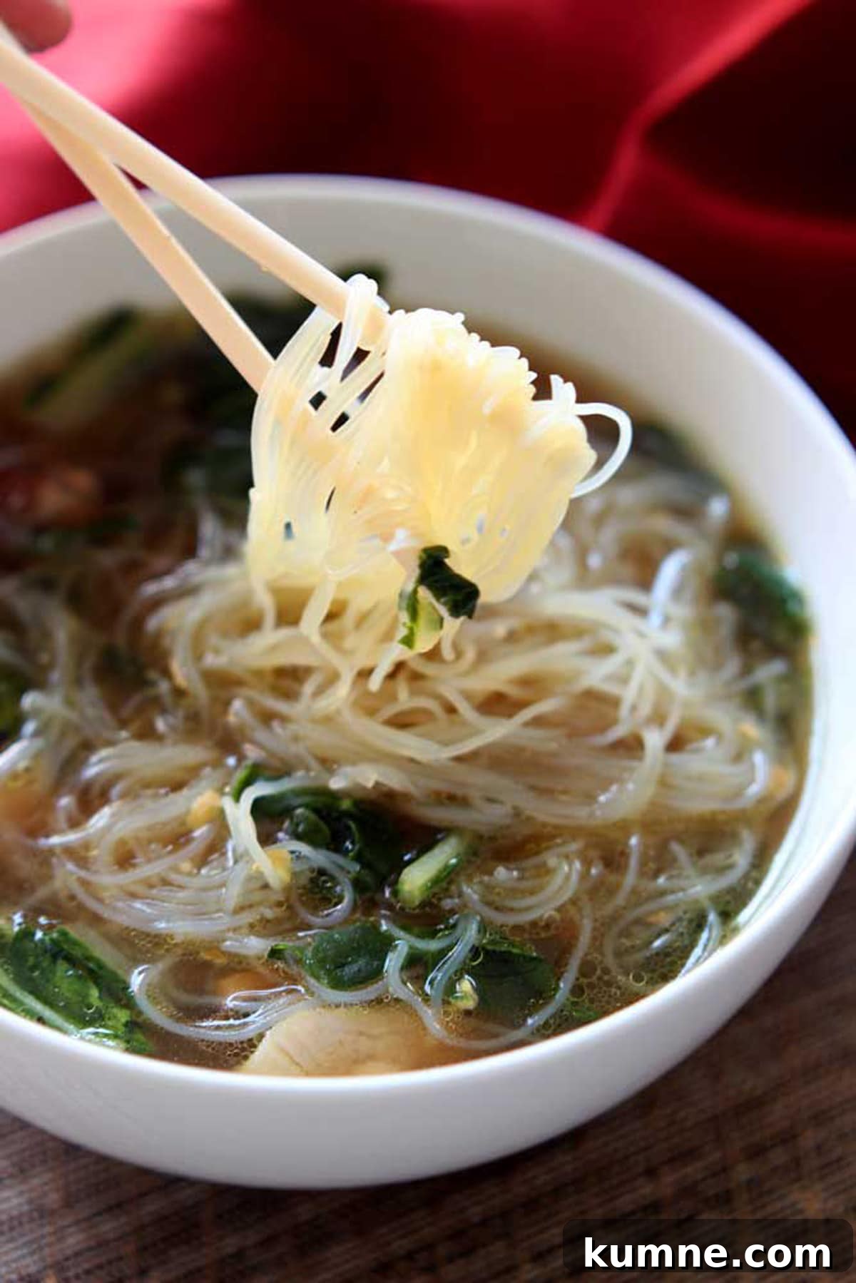 Authentic Asian Chicken Noodle Soup Recipe for Comforting Flavor 12 White bowl containing Asian Chicken Noodle Soup with Mei Fun noodles on a chopstick, red napkin in background.