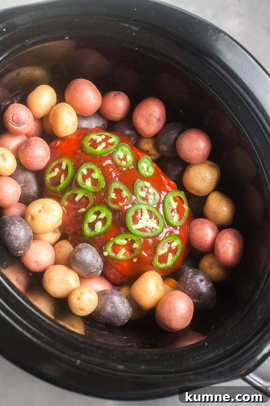 overhead view of the meatloaf and potatoes in a crock pot slow cooker. The meatloaf in the the center and is topped with homemade barbecue sauce and slices chile peppers. Small potatoes surround the meatloaf.