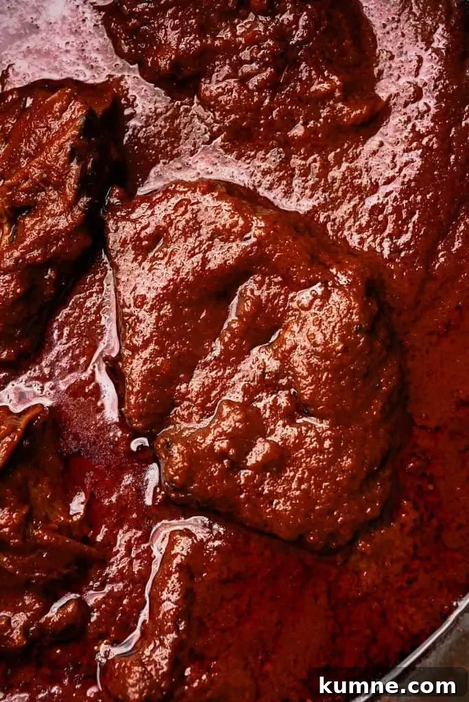 beef birria Close-up of tender beef birria simmering in a deep red chili-based sauce with visible oil and spices.