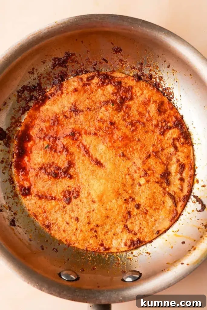 what are birria tacos Corn tortilla dipped in birria consommé frying in a skillet with a rich red chile oil coating the surface.