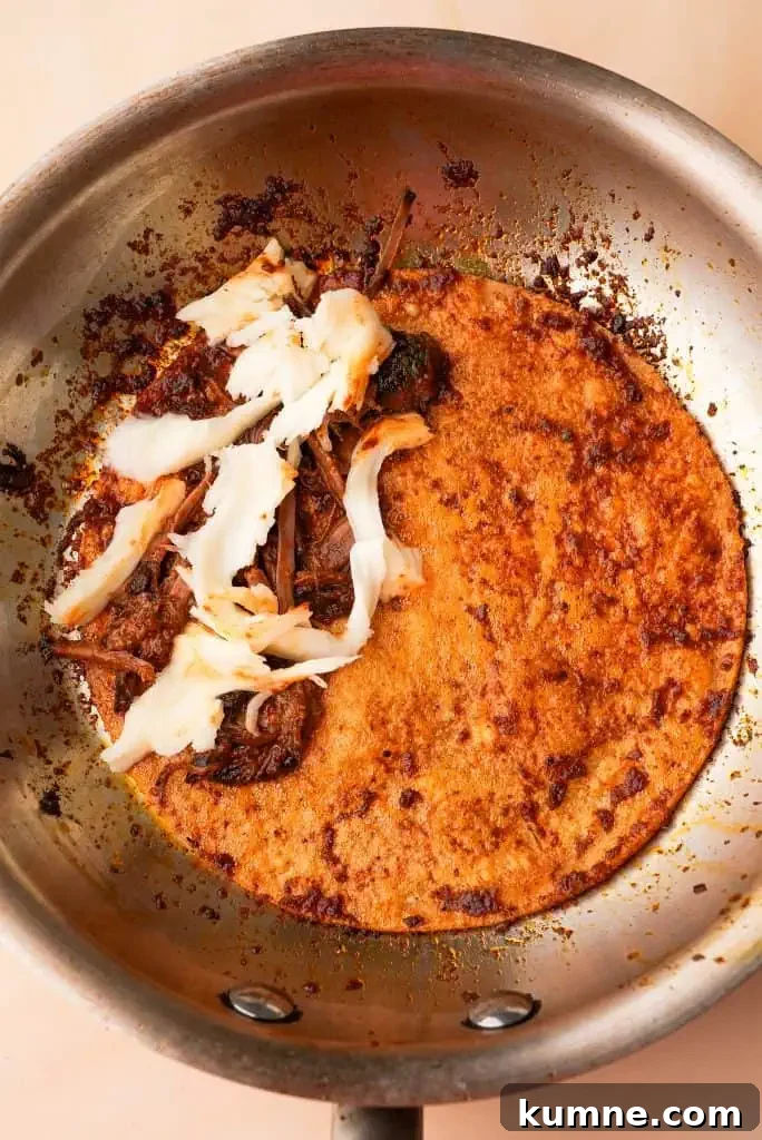 how to make birria tacos Corn tortilla in a skillet filled with shredded birria beef and melting cheese, partially folded as it cooks.