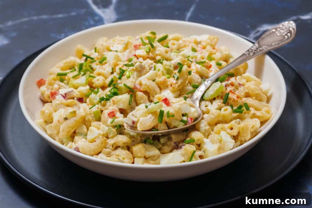 A bowl of Deviled Egg Pasta Salad with chopped vegetables and herbs, is set on a black plate, and a spoon rests in it.