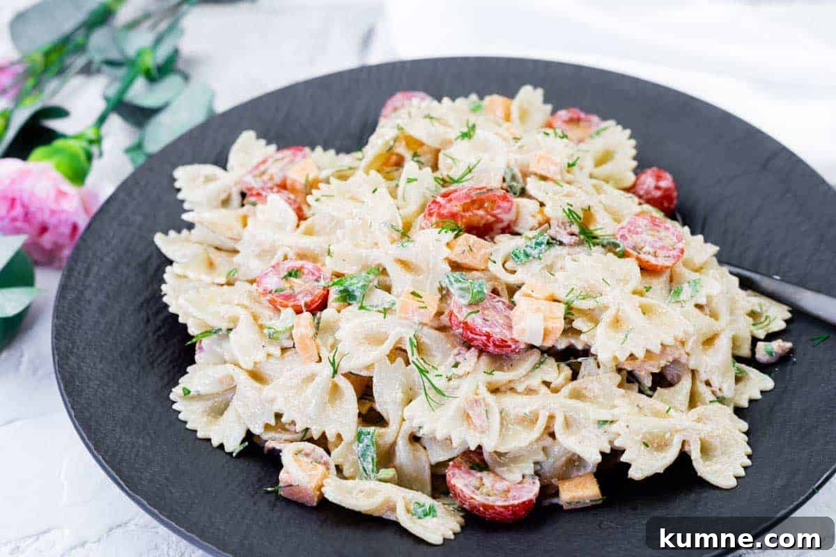 A black plate with bow-tie pasta salad mixed with cherry tomatoes, diced vegetables, herbs, and a creamy dressing.