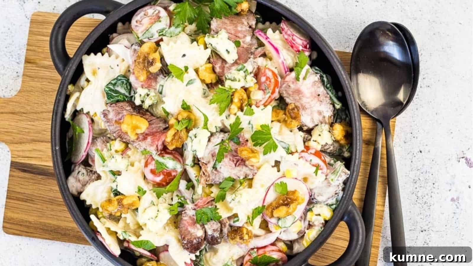 A black bowl filled with a pasta salad containing farfalle, steak slices, radishes, cherry tomatoes, lettuce, nuts, and crumbled blue cheese, garnished with parsley, on a wooden board next to a spoon.