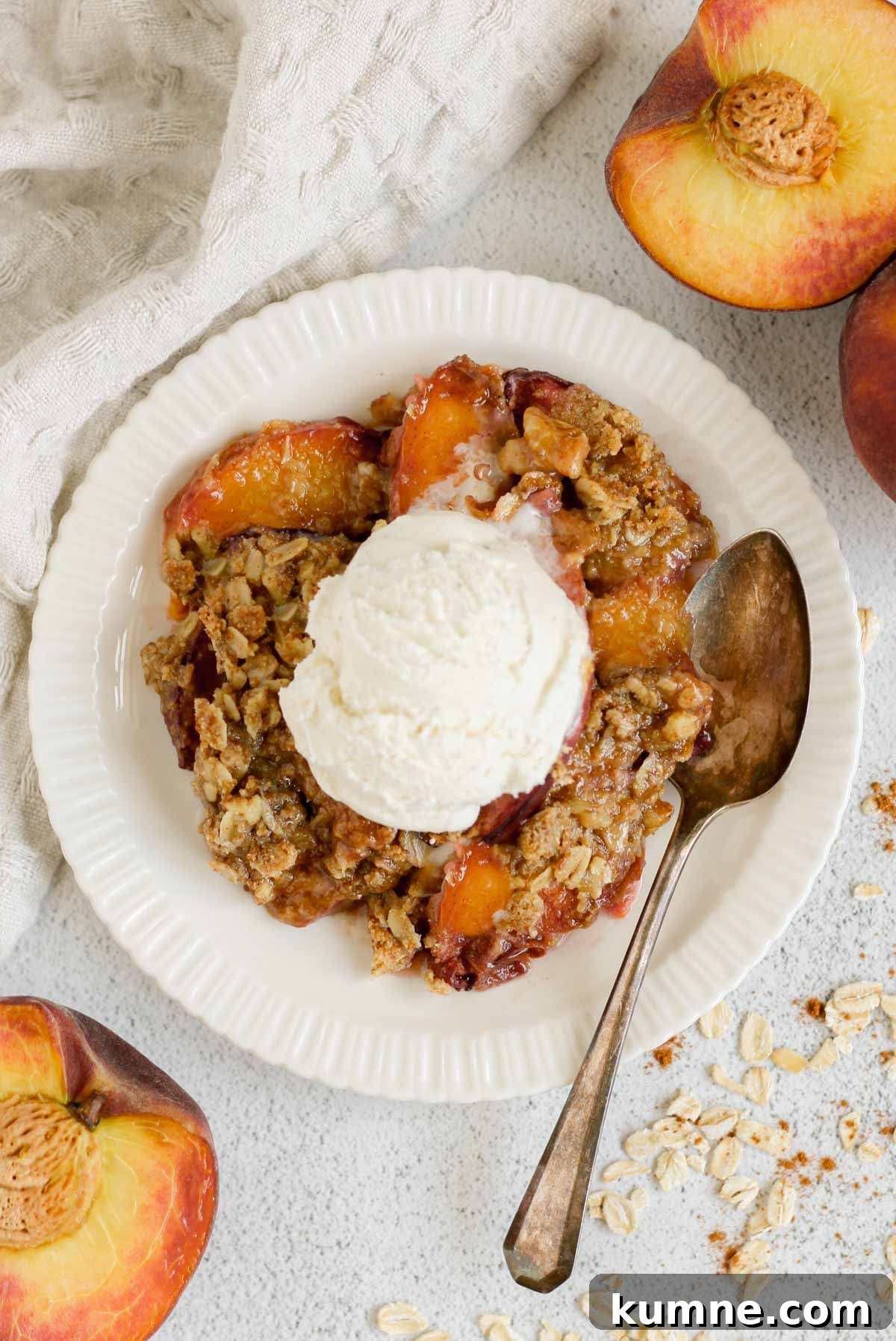 Whole Grain Low-Sugar Peach Crisp Recipe 2 Serving of peach crisp on a round plate topped with ice cream and a vintage spoon on the plate with sliced peaches on the sides of the plate.