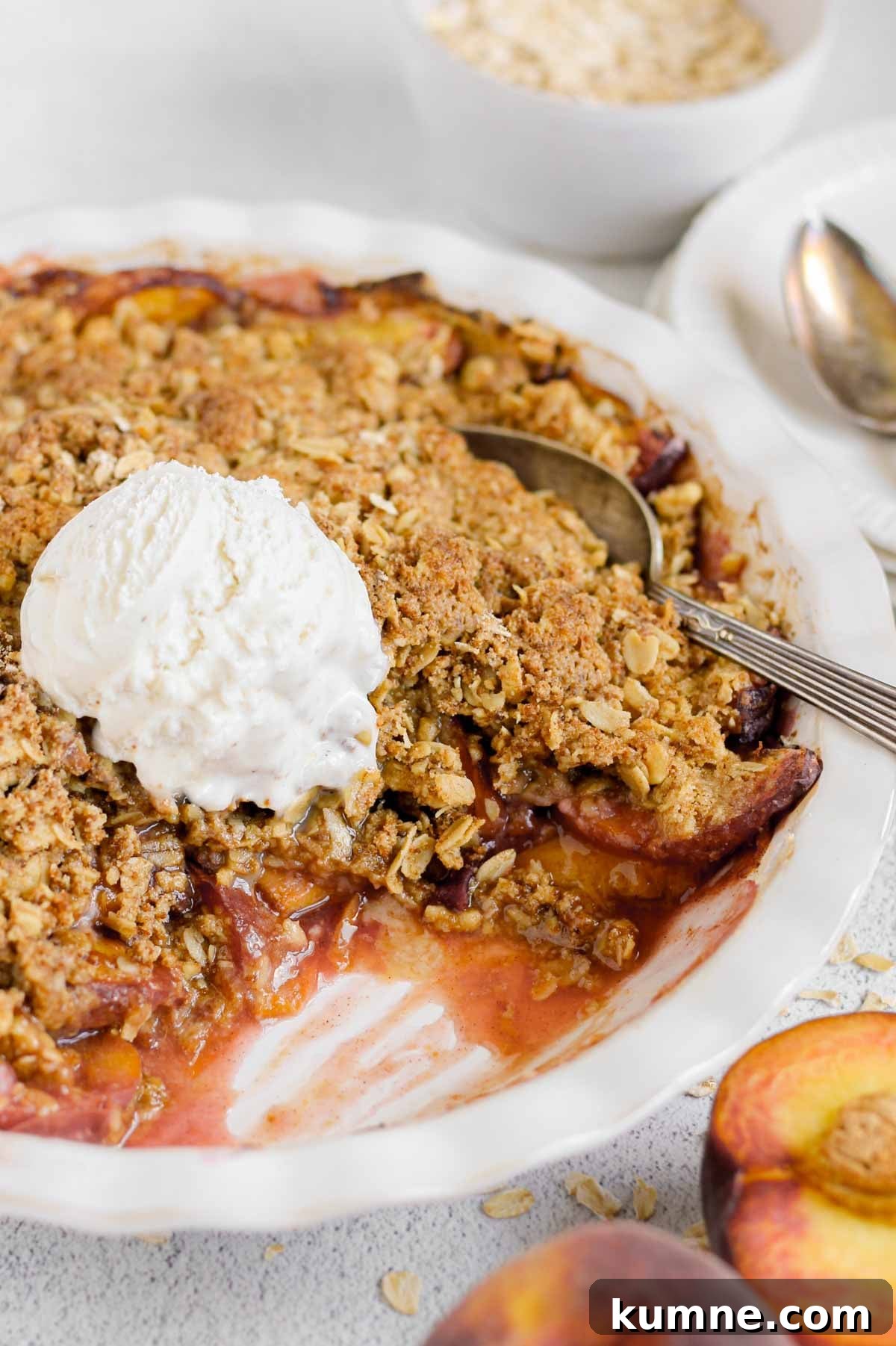 Whole Grain Low-Sugar Peach Crisp Recipe 11 A baked peach crisp in a white baking dish topped with a scoop of vanilla ice cream. A serving has been scooped out, revealing juicy peaches beneath a golden oat crumble topping. Fresh peach halves and scattered oats sit nearby.