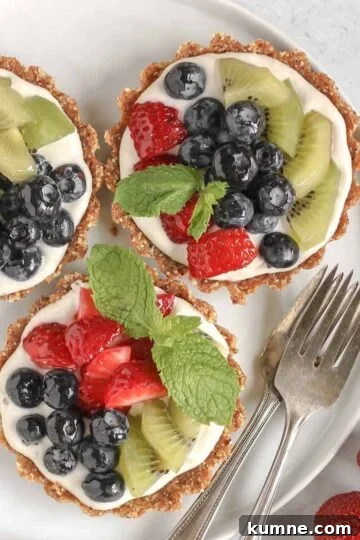 Three mini fruit tarts on a small white plate with 2 vintage forks on the plate and fresh berries off to the side.
