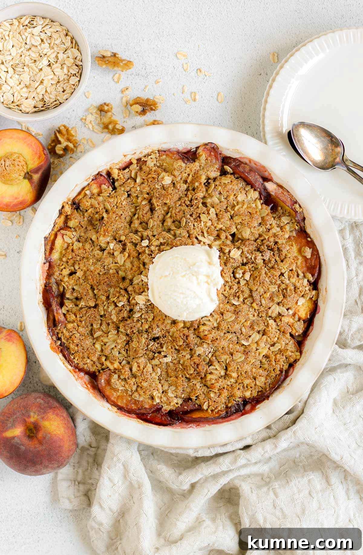 Whole Grain Low-Sugar Peach Crisp Recipe 10 Baked peach crisp topped with a scoop of ice cream in a ceramic pie dish with sliced peaches, plates, and a bowl of oats scattered on the sides.