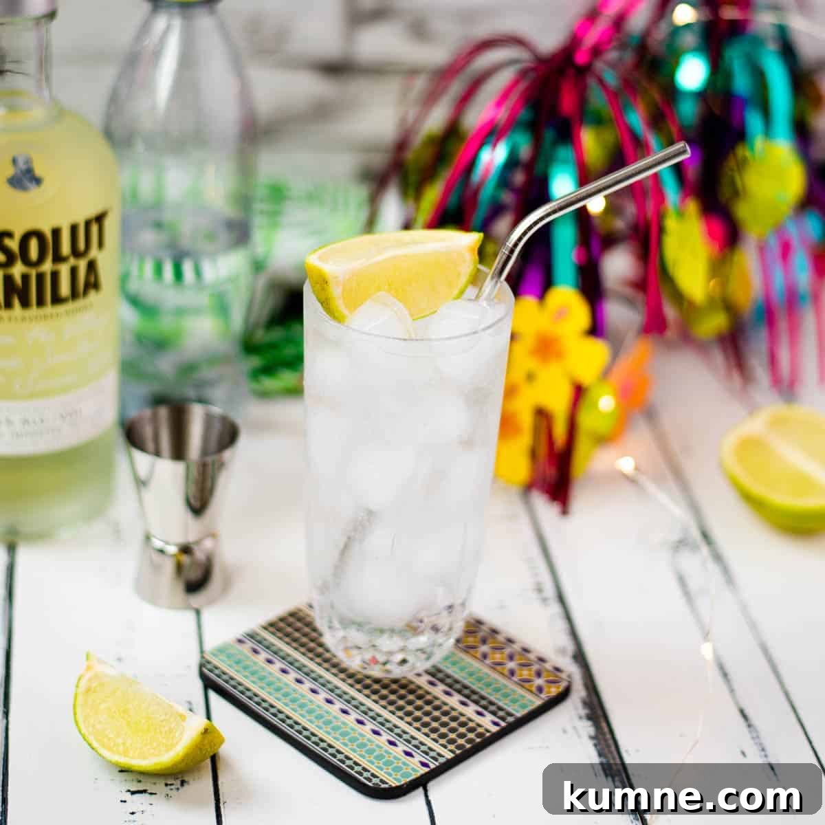 Beautiful glass filled with vanilla vodka tonic, on colorful coaster placed on white wooden board.