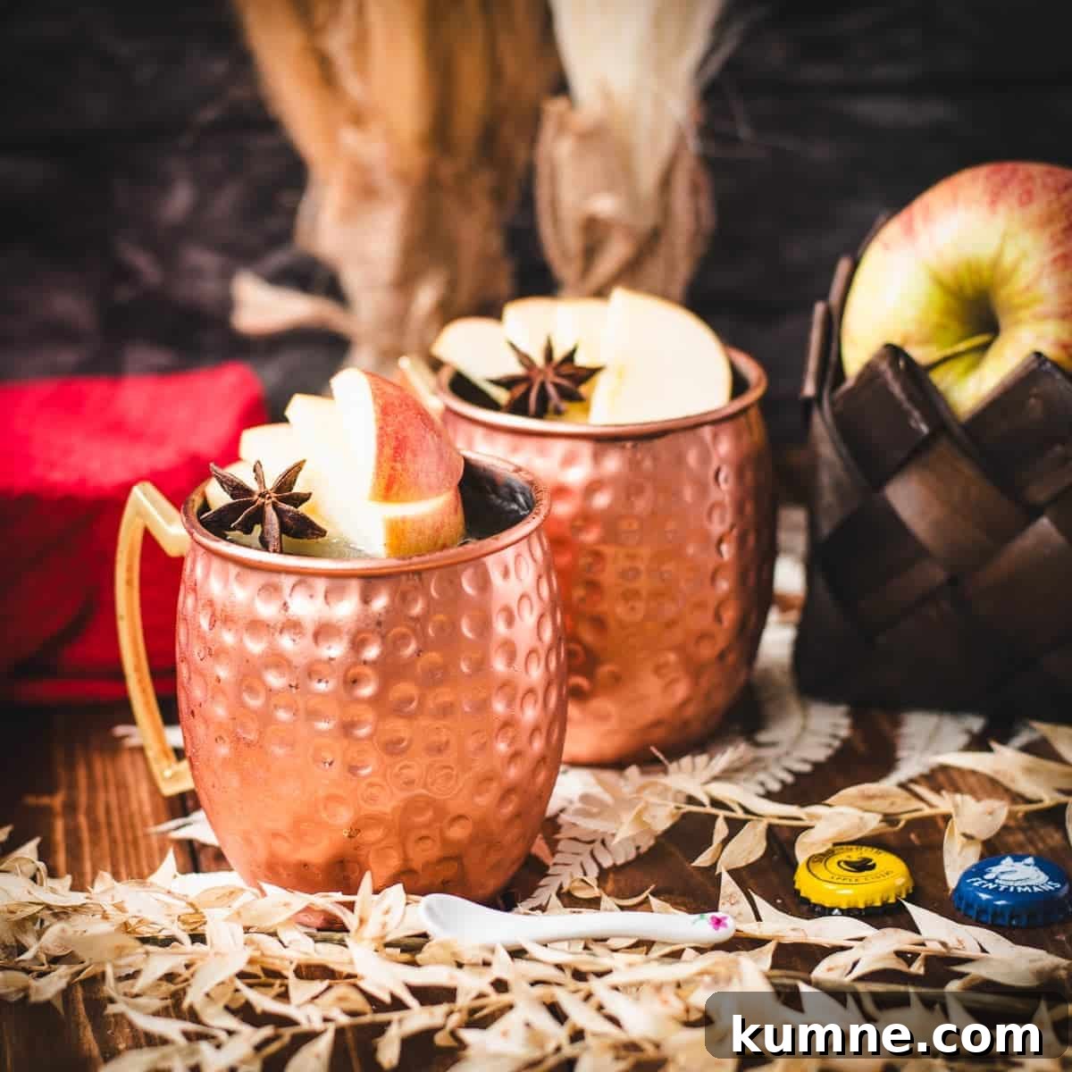 Copper mug, filled with delicious apple cider Moscow mule, garnished with fresh apple slices and a star anise. Placed on wooden board, next to a red cloth.