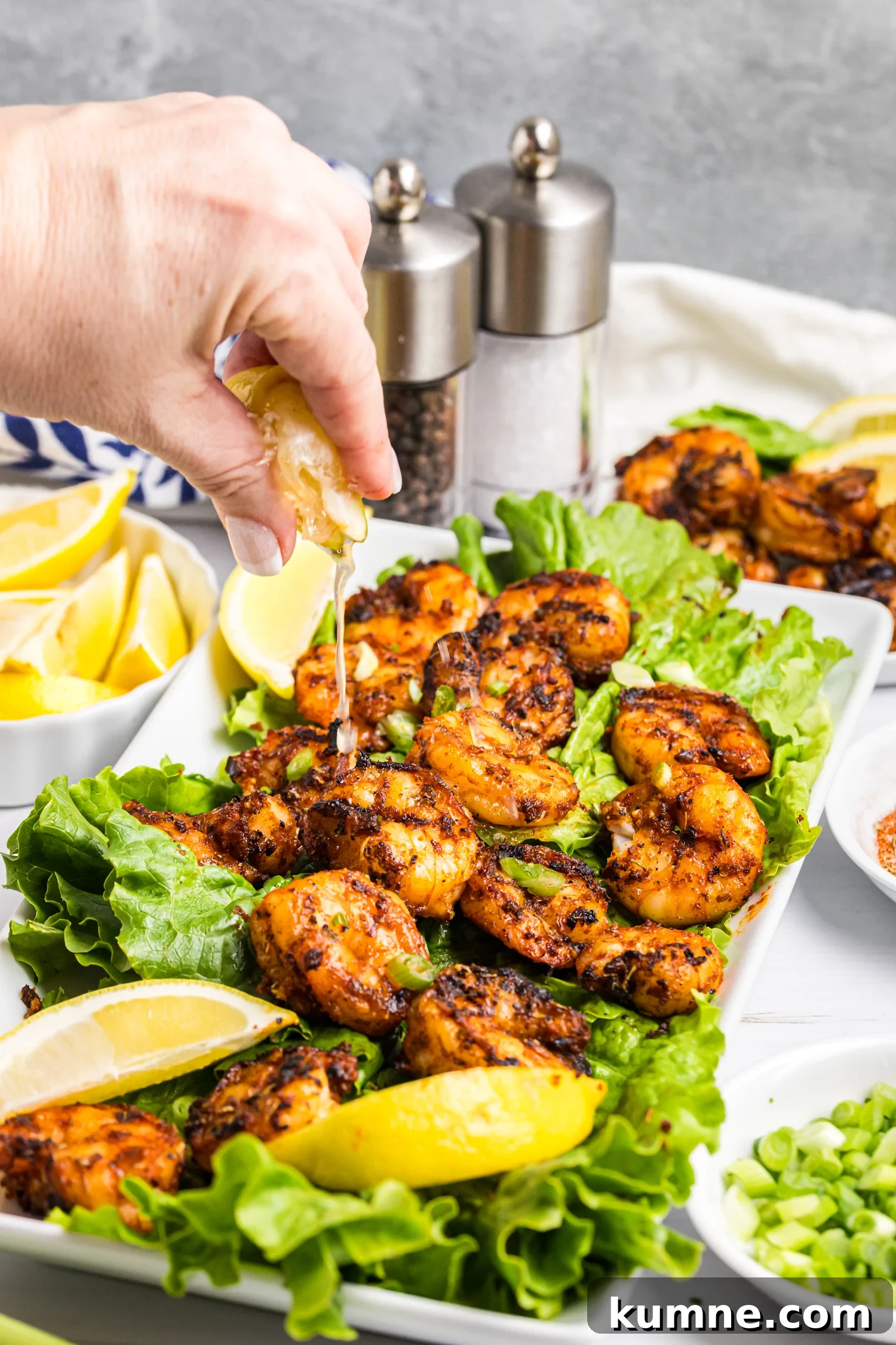Hand squeezing lemon juice over grilled shrimp served on lettuce with lemon wedges and green onions.