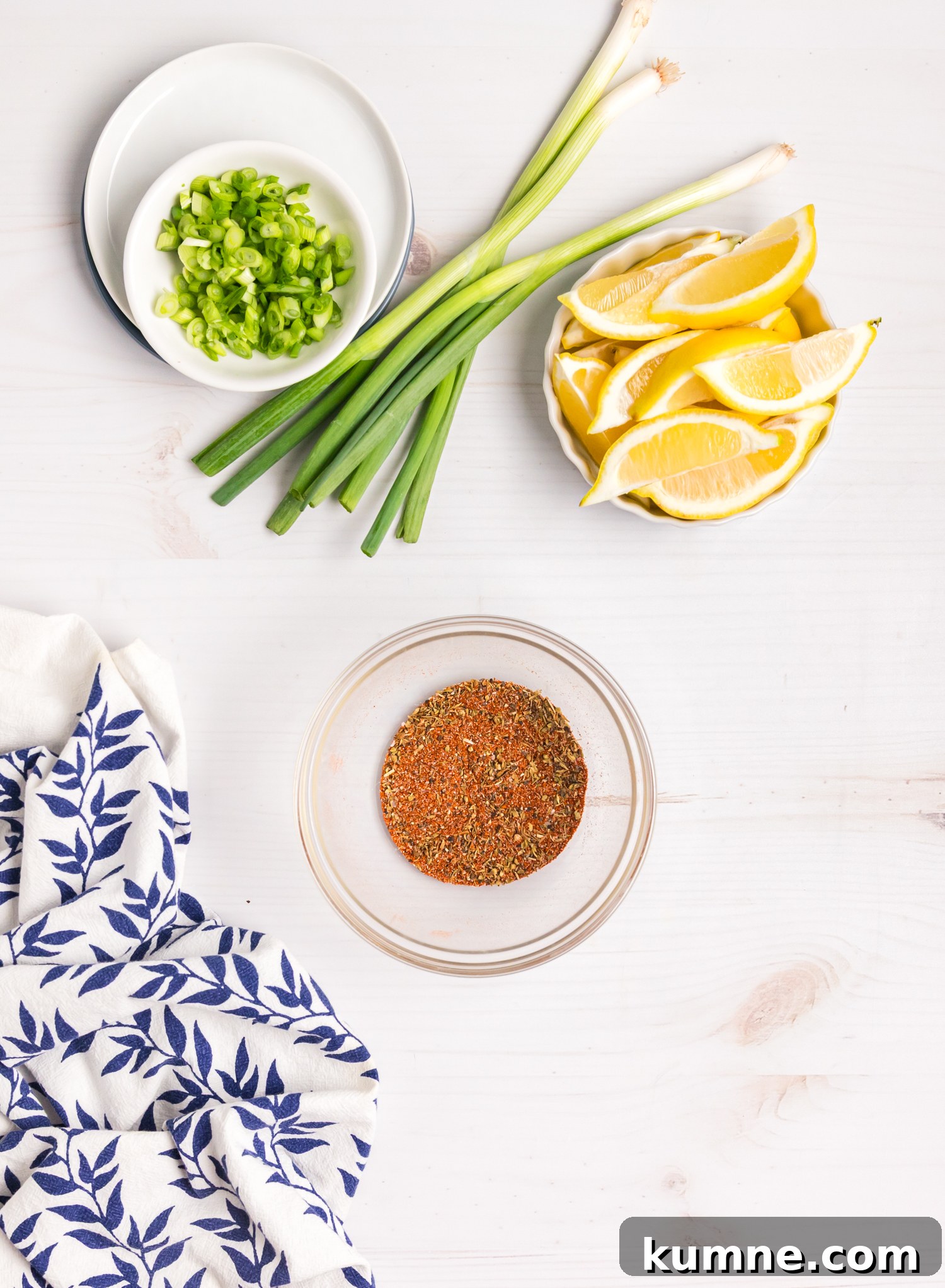 Chopped green onions, whole scallions, lemon wedges, and a bowl of spice blend arranged on a white surface.