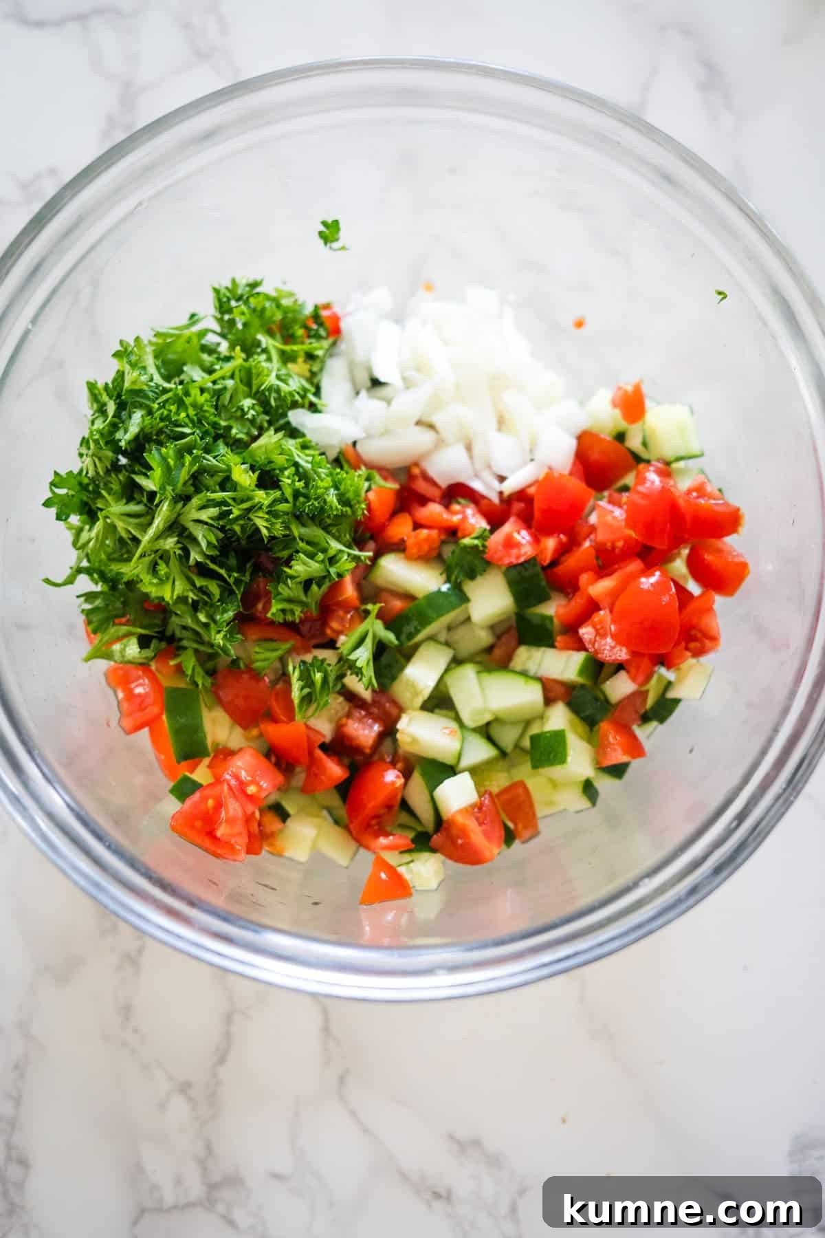 A glass bowl containing chopped tomatoes, cucumbers, onions, parsley, and tahini on a marble surface.