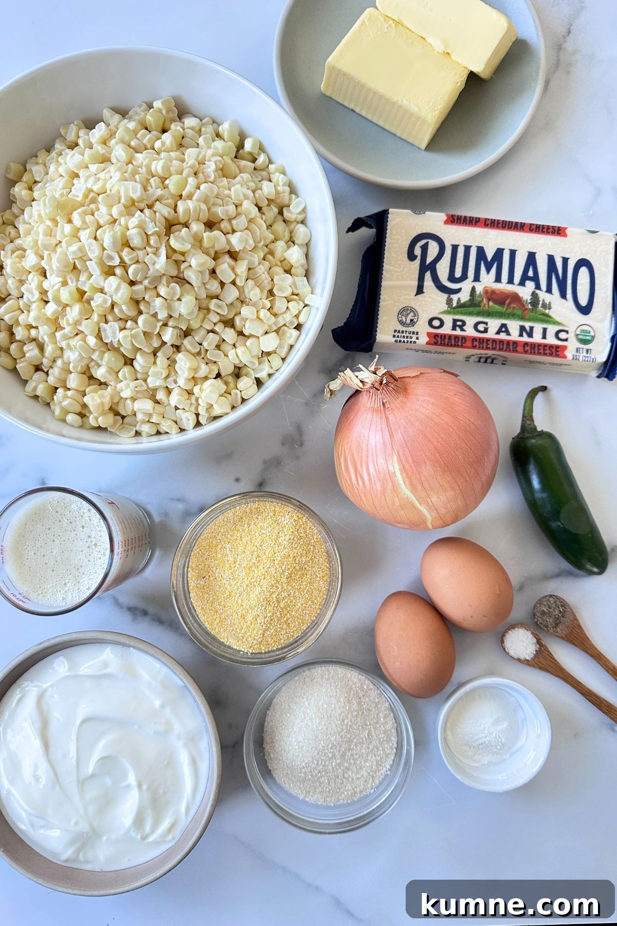 Creamy Baked Jalapeño Cheddar Corn Pudding Recipe from Scratch 2 Ingredients for corn pudding on a white counter top including corn, butter, organic Rumiano sharp cheddar cheese, onion, jalapeno, eggs, spices, Greek yogurt, flour, and cornmeal.