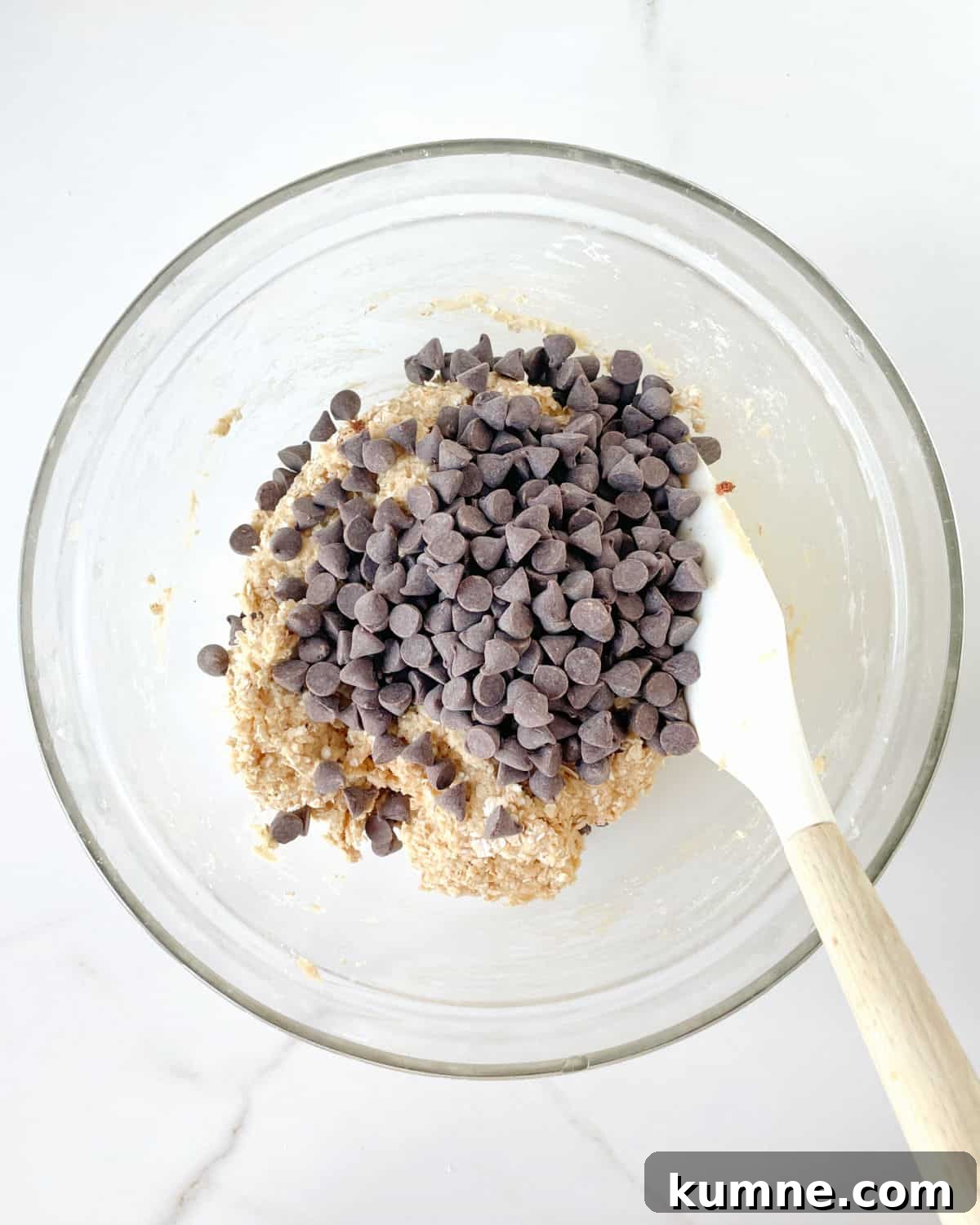 Chewy Chocolate Chip Oatmeal Cookies Recipe for Homemade Treats 5 Chocolate chips added to cookie dough in a glass bowl with a white silicon spatula. White marble surface.