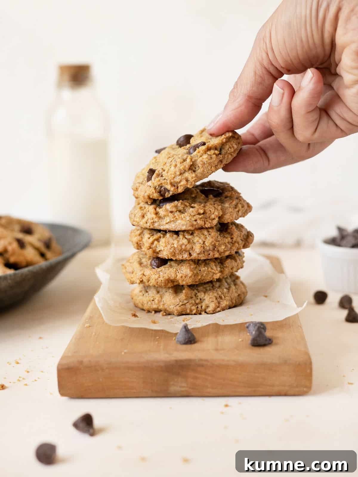 Chewy Chocolate Chip Oatmeal Cookies Recipe for Homemade Treats 9 Hand lifting chocolate chip oatmeal cookie from a stack on a wooden board. Milk bottle and chips in the white background.