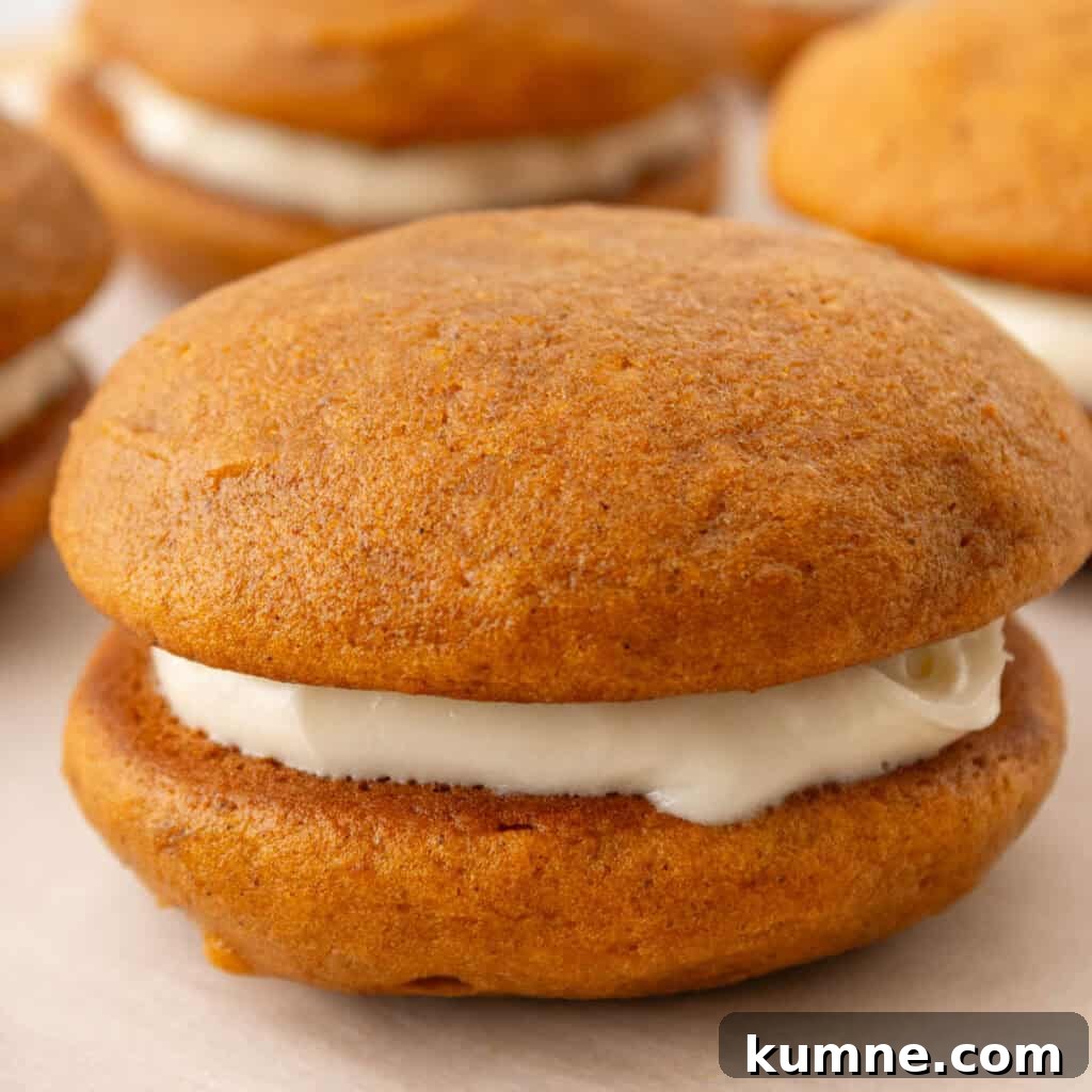 Close-up image of a pumpkin whoopie pie with a creamy filling, resting on a white surface. Other whoopie pies are blurred in the background.