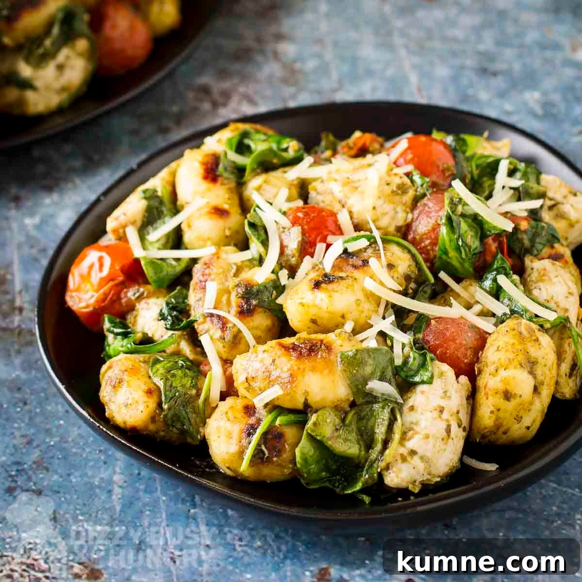 Side shot of two plates of pesto chicken and gnocchi sprinkled with shredded parmesan on black plates.