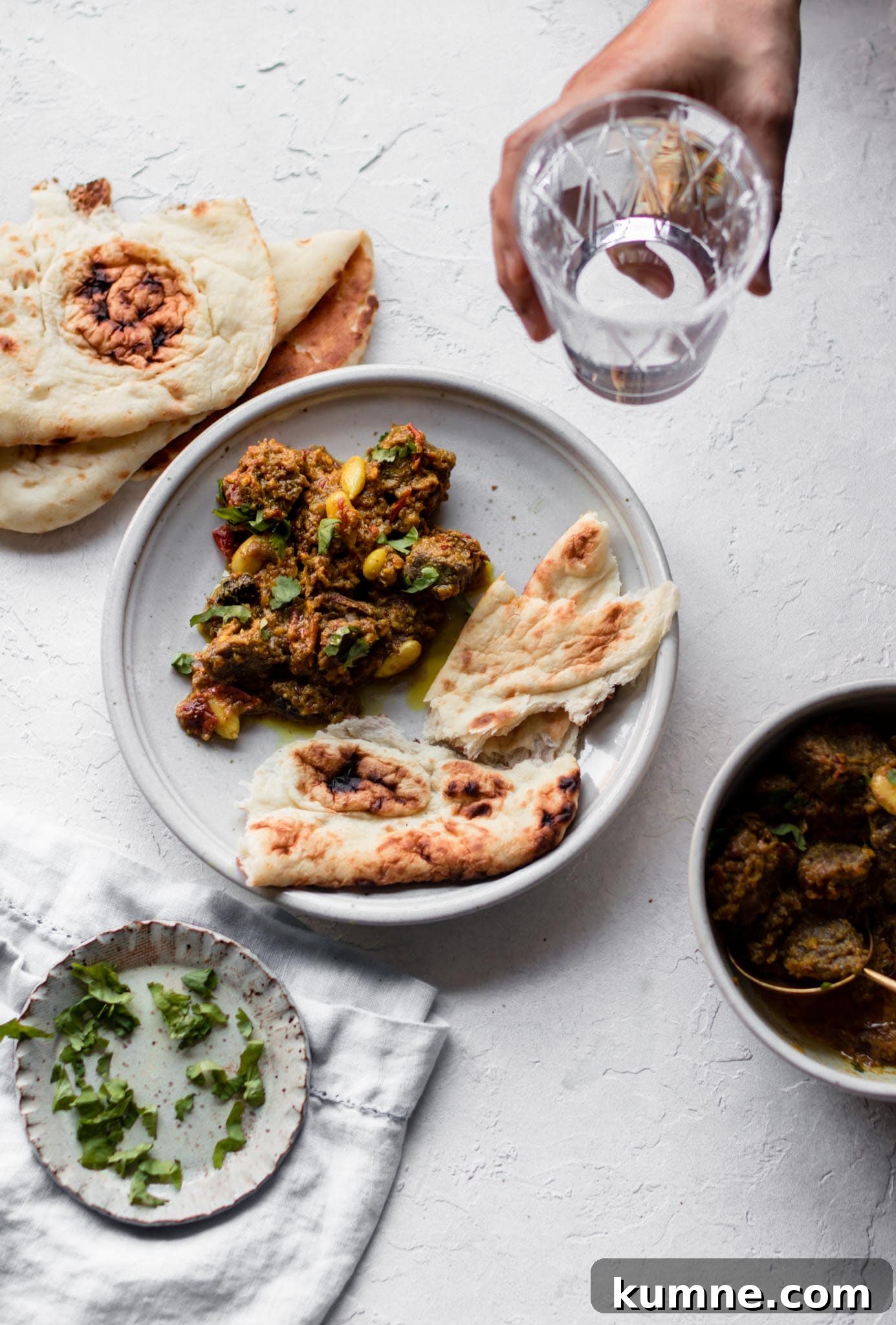 Picking up a glass of water above Pakistani-style Beef Curry (Stew) on a plate with naan