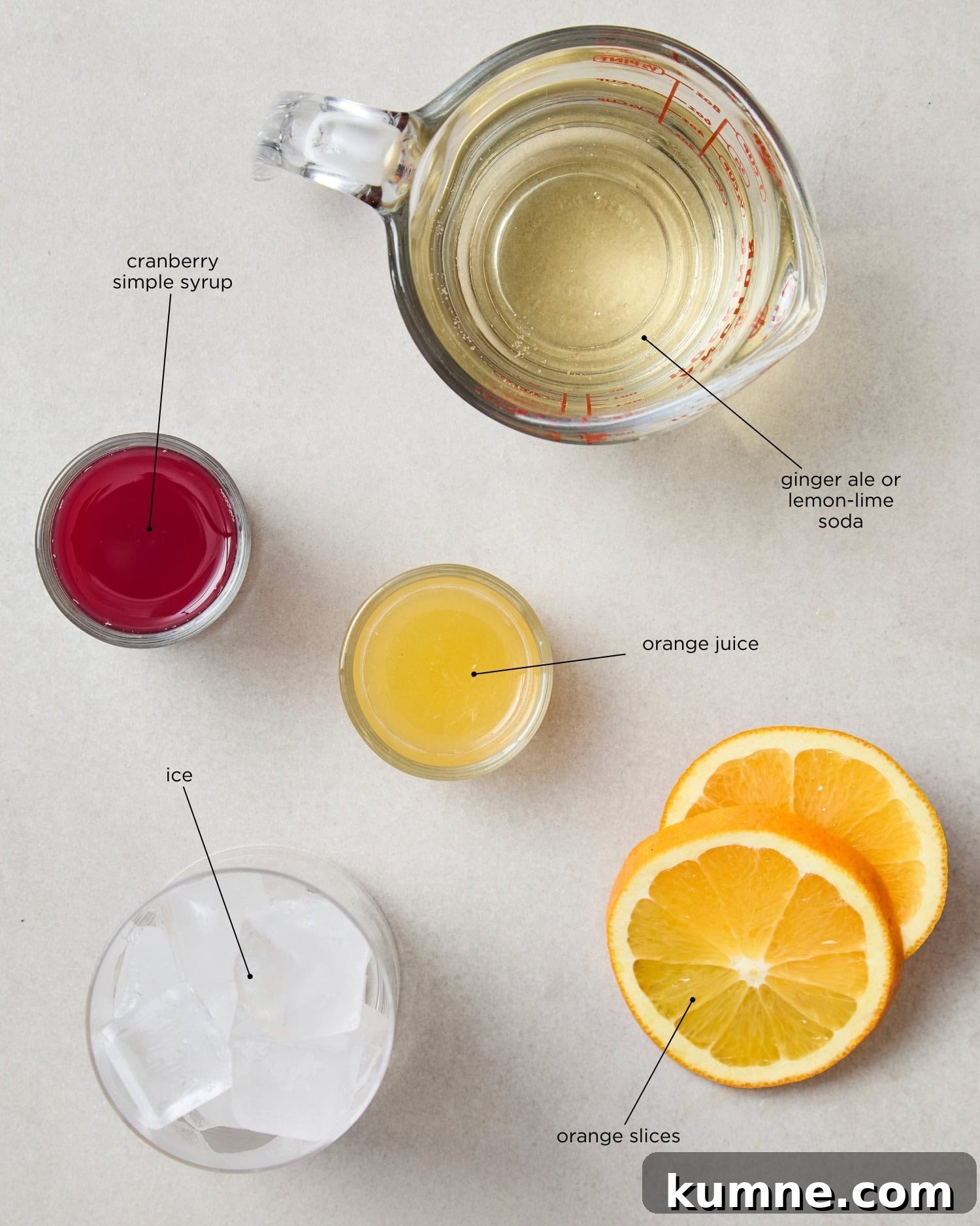 Concrete surface with ingredients for a cranberry mocktail including simple syrup, orange juice, ice, and more.