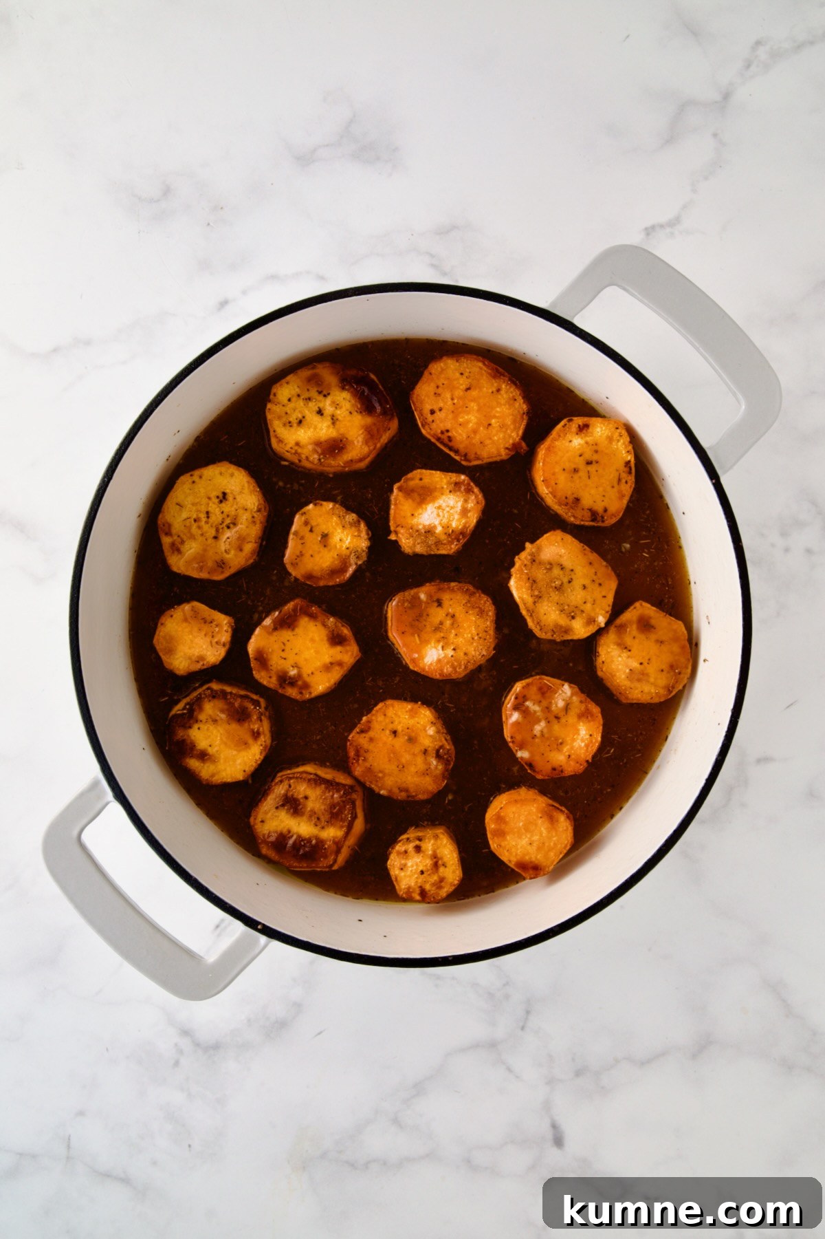 Rounds of browned sweet potatoes in white enamel cast iron skillet in herb and broth blend.