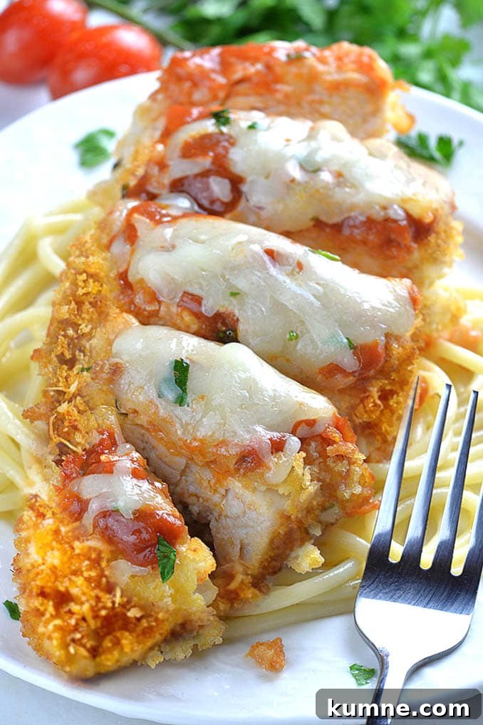 Whole piece of Baked Chicken Parmesan on a white plate laying on spaghetti with fork.