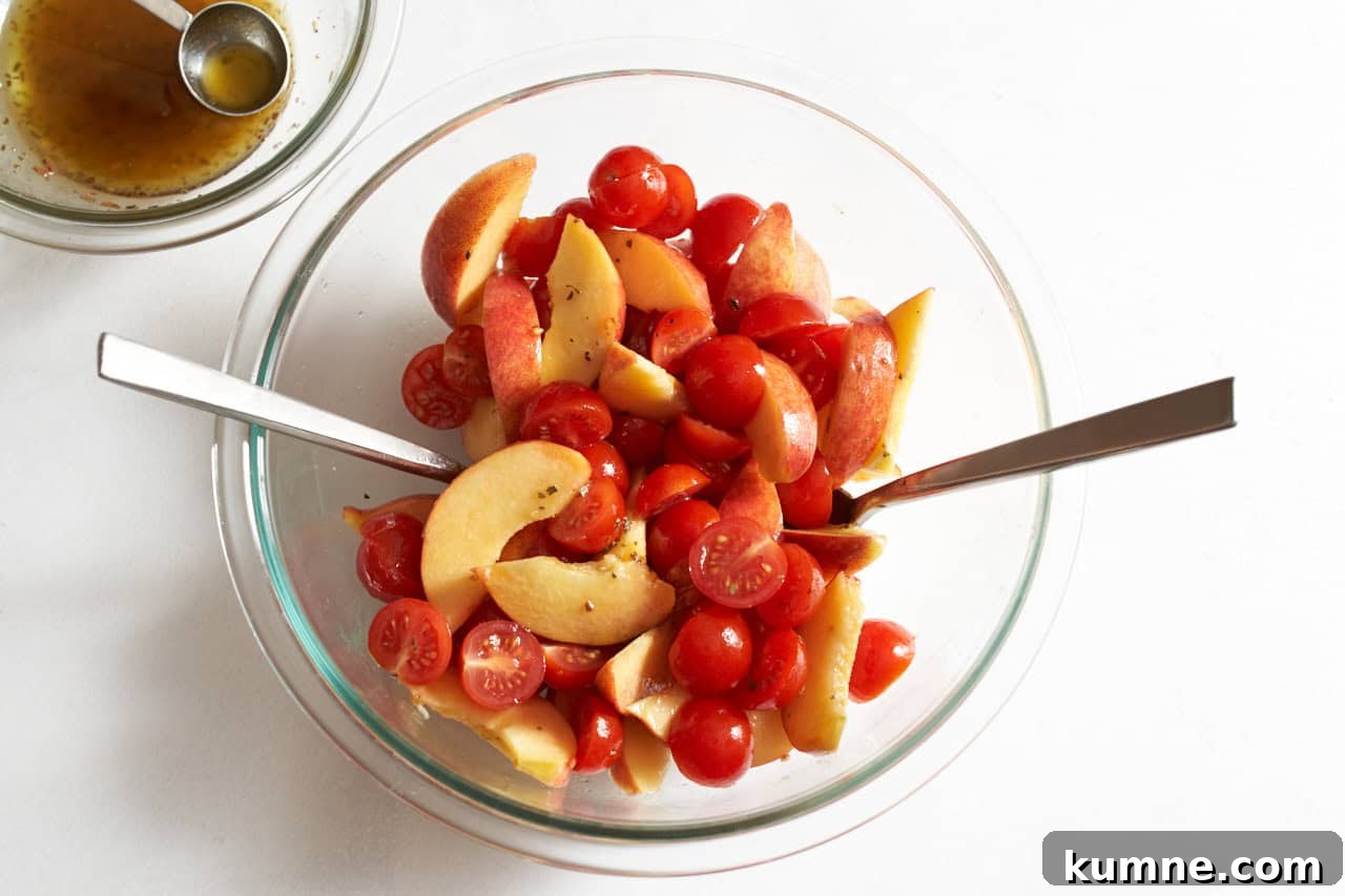 A bowl of sliced peaches and cherry tomatoes tossed in Italian dressing, with two spoons in it.