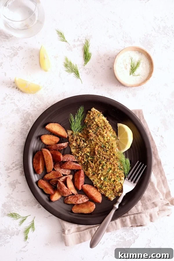 Photo of Pistachio-Crusted Fish with Lemon-Dill Aioli served with roasted potatoes garnished with lemon slices and dill sprigs on black plate.